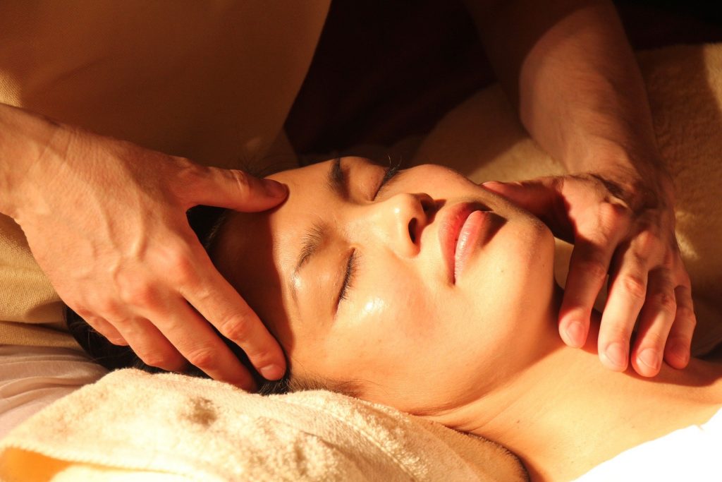 Visage d'une femme asiatique allongée sur une table de massage, yeux fermés, deux mains du praticien, une sur son cou, une sur son front, pour traiter les maux et douleurs dont elle souffre par le biais de la médecine traditionnelle chinoise.