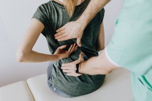 Traitement ostéopathique du mal de dos d'une femme en tee-shirt gris foncé, de dos. Un professionnel de santé a ses mains appliqués sur son dos pour la soulager